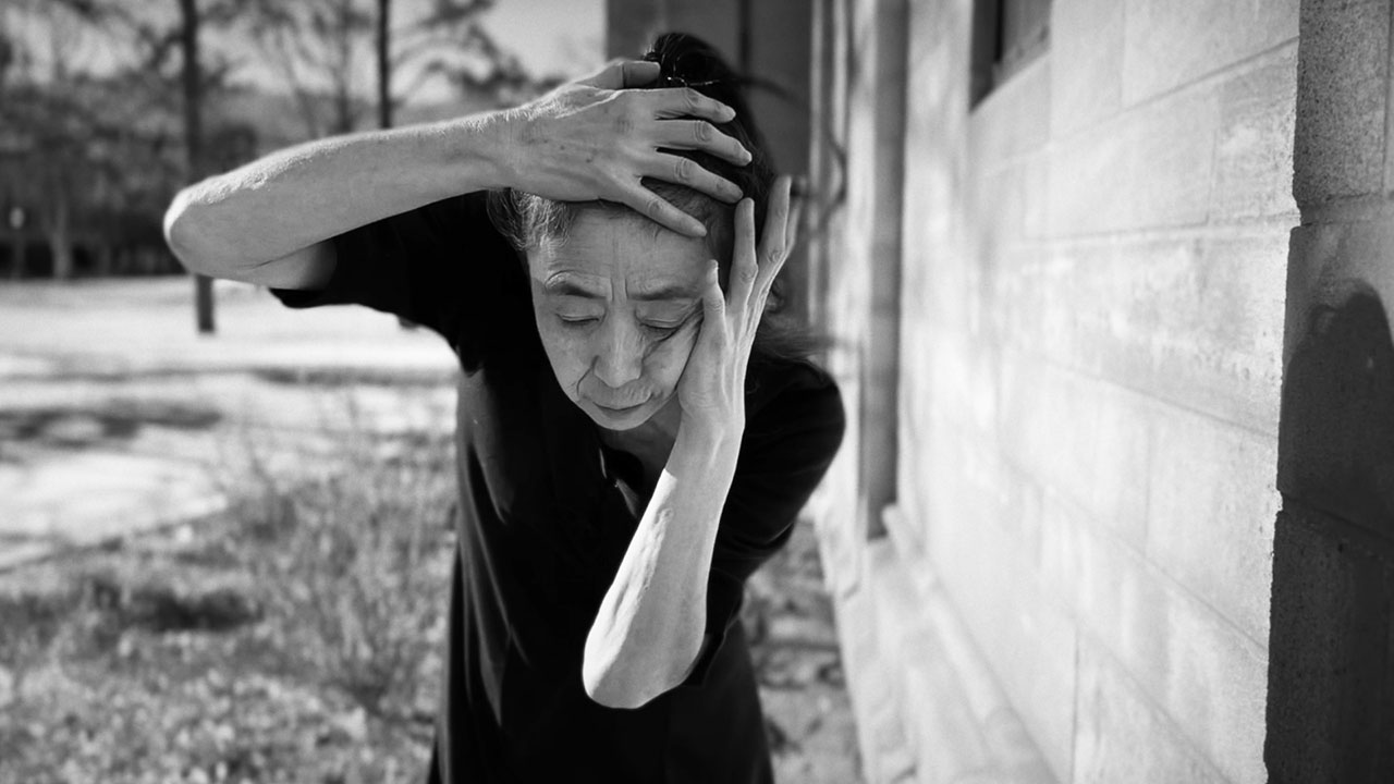 An Asian woman stands outside in the sun. She is bending forward slightly, with one hand on her cheek and one on her forehead, her eyes down.