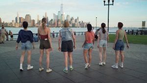 A group of tennis-shoe clad dancers stands in a park looking out at the New York City skyline. Their backs are to the camera and they wear shorts with colorful tops.