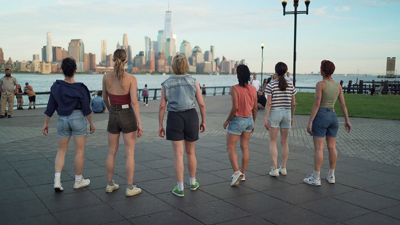 A group of tennis-shoe clad dancers stands in a park looking out at the New York City skyline. Their backs are to the camera and they wear shorts with colorful tops.