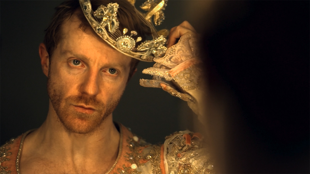 Steven McCrae is a male ballet dancer with grey eyes, red hair, and a short beard. He stares intently at his reflection in a mirror while removing a crown from atop his head. He wears a peach-colored ballet costume with intricate beading.