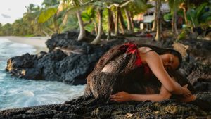 Two Fijian dancers sit huddled in an embrace on the rocky shoreline of a tropical beach. The upper dancer curls over the lower dancer and wears a red feathered top and grass skirt. In the background are palm trees and a beach.
