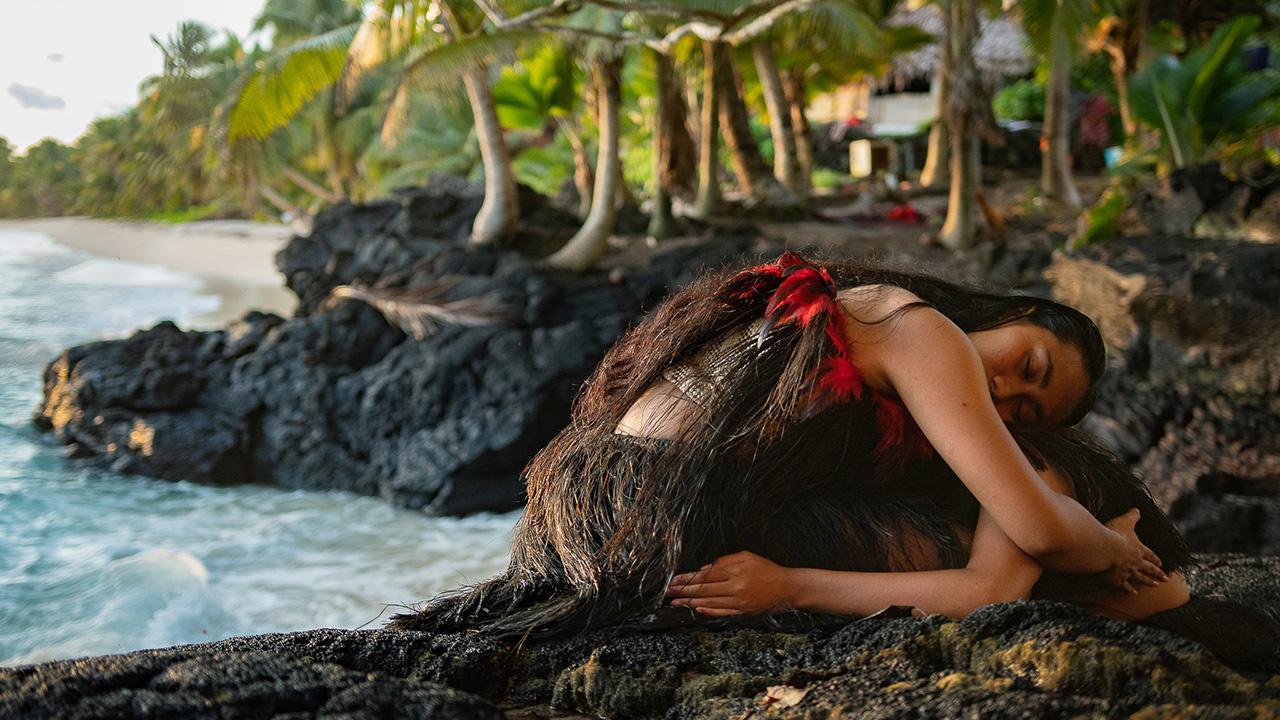 Two Fijian dancers sit huddled in an embrace on the rocky shoreline of a tropical beach. The upper dancer curls over the lower dancer and wears a red feathered top and grass skirt. In the background are palm trees and a beach.