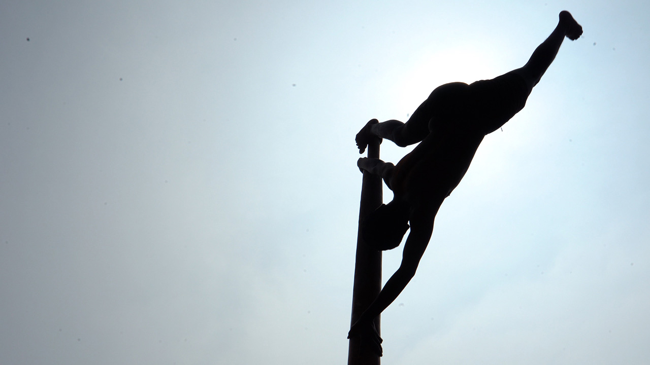 View from below of a silhouetted dancer hanging upside down on a large pole. He holds the pole with both arms, one leg resting above his hands and one leg outstretched to the side in a split in the air. The sun shines brightly behind him.