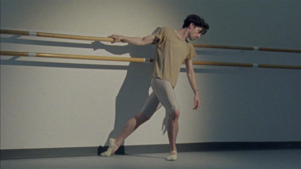 Male ballet dancer Joseph Walsh poses at a ballet barre. He leans, hanging onto the barre with one hand, and looks downwards. He wears a baggy beige t-shirt, silver tights, and light beige ballet shoes.