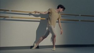 Male ballet dancer Joseph Walsh poses at a ballet barre. He leans, hanging onto the barre with one hand, and looks downwards. He wears a baggy beige t-shirt, silver tights, and light beige ballet shoes.