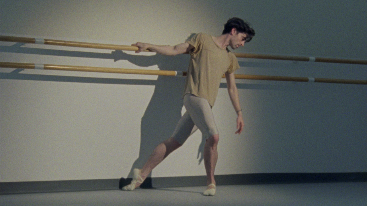 Male ballet dancer Joseph Walsh poses at a ballet barre. He leans, hanging onto the barre with one hand, and looks downwards. He wears a baggy beige t-shirt, silver tights, and light beige ballet shoes.
