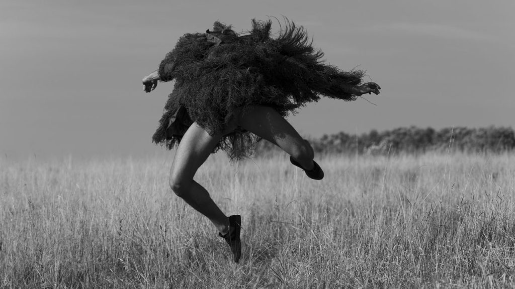 A person jumps in a wheat field with their arms extended and their head leaning back out of view. They wear a black, stringy, long-sleeved dress-like garment resembling animal hair, and black sneakers.