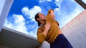 A dancer in a yellow knitted top leans to the side slightly, her hands in front of her chest, elbows out to the side and her eyes closed. She stands outside in between cement walls, and in the sky behind her puffy white clouds.