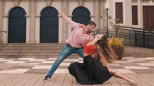 A man in a pink shirt and jeans holds a woman in a red tank top and flowing back skirt close to the ground in a dip. His free arm extends up and to the side, and her free arm is extended in the opposite direction, resting on her leg. She smiles up at him, her long hair flowing behind her. They stand on the sidewalk in front of a building with three blue doors that arch at the top.