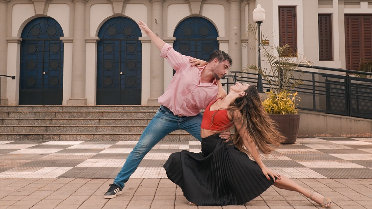 A man in a pink shirt and jeans holds a woman in a red tank top and flowing back skirt close to the ground in a dip. His free arm extends up and to the side, and her free arm is extended in the opposite direction, resting on her leg. She smiles up at him, her long hair flowing behind her. They stand on the sidewalk in front of a building with three blue doors that arch at the top.
