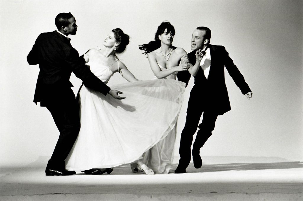 A greyscale image of two men in tuxedos alongside two women in formal gowns. The couple on the left strikes a traditional dance pose, while the couple on the right is caught mid-motion. The woman holds on the man's arm as the man move sideways, a funny expression on his face.