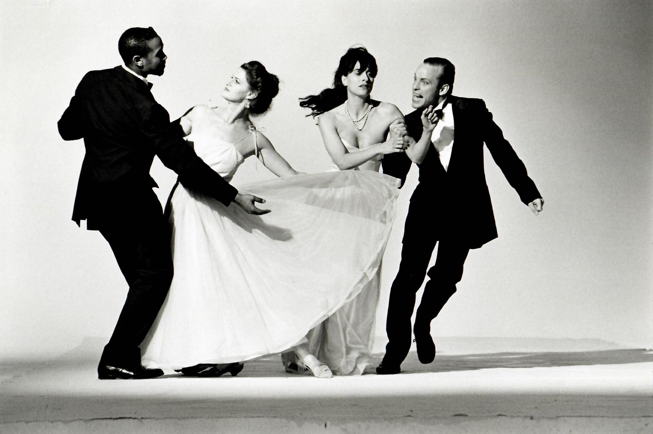 A greyscale image of two men in tuxedos alongside two women in formal gowns. The couple on the left strikes a traditional dance pose, while the couple on the right is caught mid-motion. The woman holds on the man's arm as the man move sideways, a funny expression on his face.