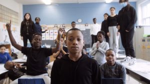 A child sits in a classroom, gazing solemnly into the camera lens. Behind him are his classmates standing on and sitting at desks. On holds his arms to his sides, index fingers pointed upwards. Another has his hands together in a praying position, eyes closed.