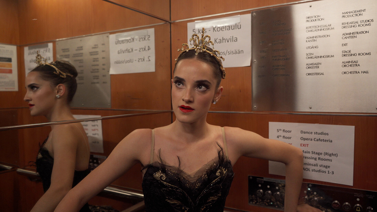 A dancer in stage makeup, a golden tiara and a black feathered costume looks up. She stands in an elevator, and her reflection can be seen behind her. She holds her arms on the railings on the sides of the elevator.