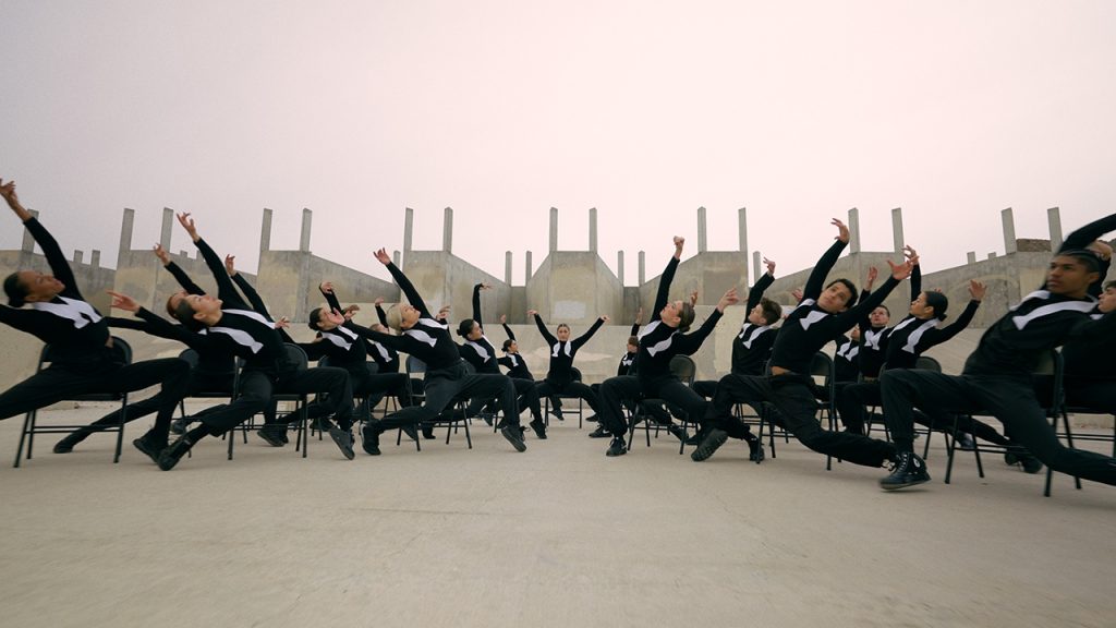 A large group of dancers are posed in black chairs in a vast industrial lot. They lean right and left, parted in the middle by a dancer who faces the camera straight on with their arms raised. The dancers are uniformly dressed in black long-sleeve shirts, black pants, and black shoes, with a white untied bowtie around their necks.