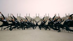 A large group of dancers are posed in black chairs in a vast industrial lot. They lean right and left, parted in the middle by a dancer who faces the camera straight on with their arms raised. The dancers are uniformly dressed in black long-sleeve shirts, black pants, and black shoes, with a white untied bowtie around their necks.