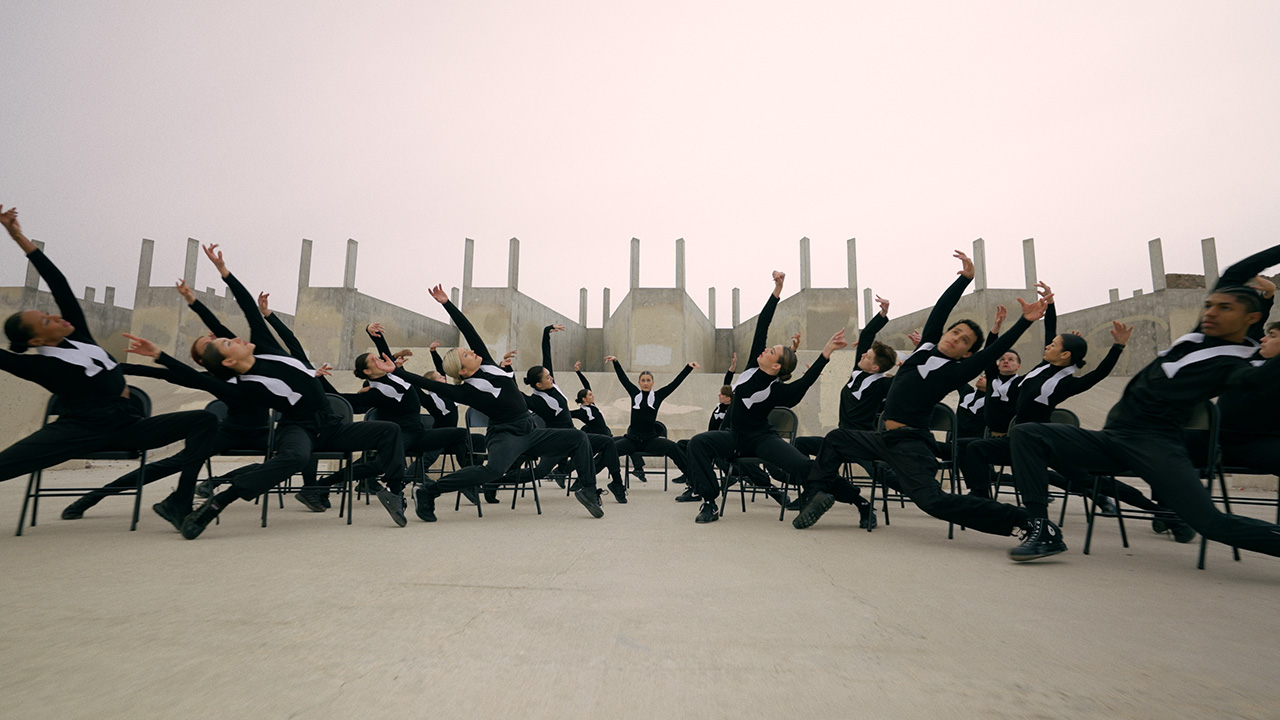 A large group of dancers are posed in black chairs in a vast industrial lot. They lean right and left, parted in the middle by a dancer who faces the camera straight on with their arms raised. The dancers are uniformly dressed in black long-sleeve shirts, black pants, and black shoes, with a white untied bowtie around their necks.