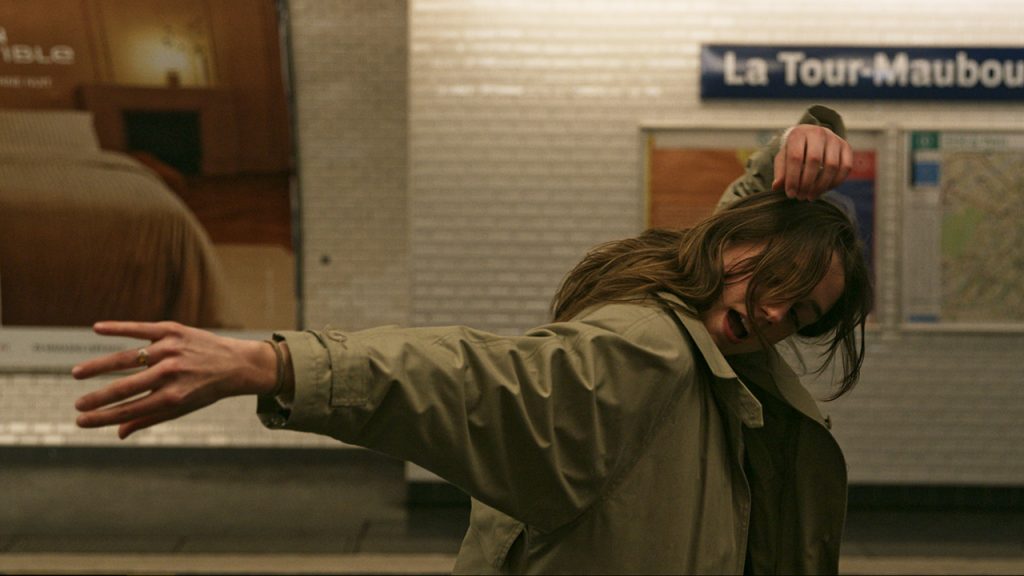 On a Parisian Métro platform, a woman twists her right shoulder forward while extending her right arm sideways. Her left arm fold above her head. She wears an army green jacket and her long brunette hair obscures most of her face.
