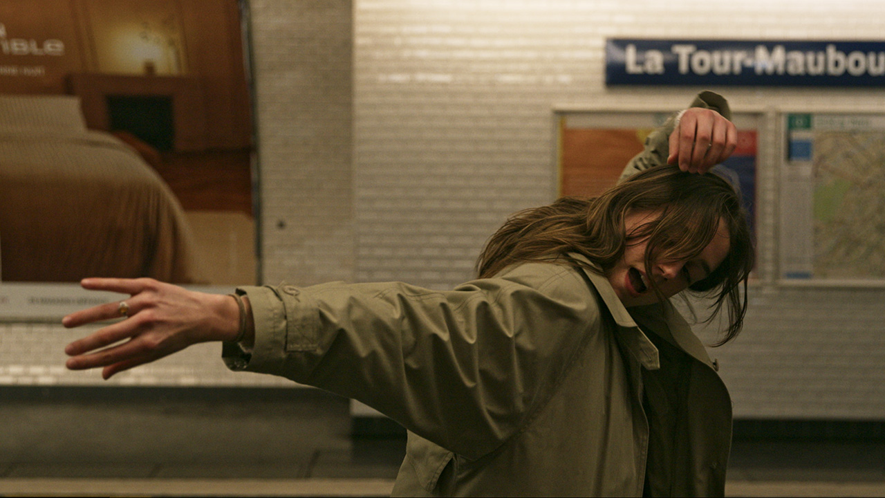 On a Parisian Métro platform, a woman twists her right shoulder forward while extending her right arm sideways. Her left arm fold above her head. She wears an army green jacket and her long brunette hair obscures most of her face.