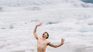 A shirtless man stands on a glacier. His arms are outstretched, hands facing outwards, and his mouth is wide open in a scream.