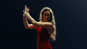 A woman looks intensely into the camera lens against a dark backdrop. She wears a bright red dress and holds her hands out to the side, raised dramatically, as if mid-clap. She has long dark blonde hair that cascades down her back.