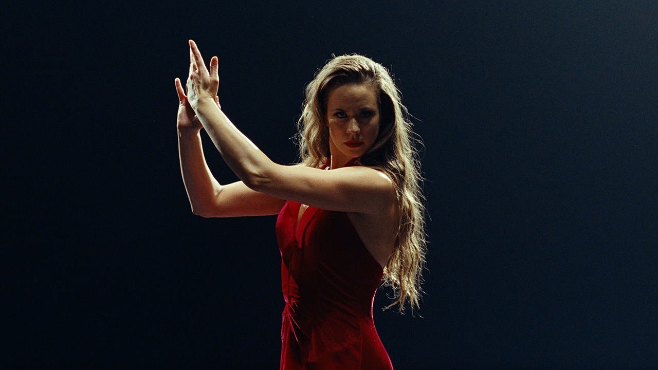 A woman looks intensely into the camera lens against a dark backdrop. She wears a bright red dress and holds her hands out to the side, raised dramatically, as if mid-clap. She has long dark blonde hair that cascades down her back.