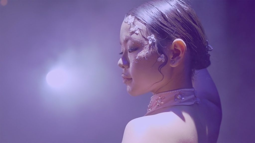 A girl with slicked back hair held by a clip looks down to her side, a bright stage light on her face. She wears a pink collar with rhinestones and pearls, and has white flowers cascading down the side of her face.