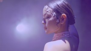A girl with slicked back hair held by a clip looks down to her side, a bright stage light on her face. She wears a pink collar with rhinestones and pearls, and has white flowers cascading down the side of her face.