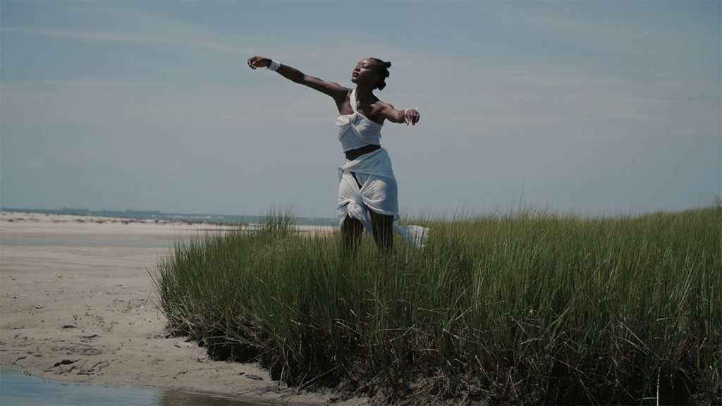 A dancer stands in tall grass along a shoreline. They wear a crop top and skirt made out of knotted white fabric and their hair forms three buns down the back of their head. They stand, extending their arms outwards, as sunlight hits their face.