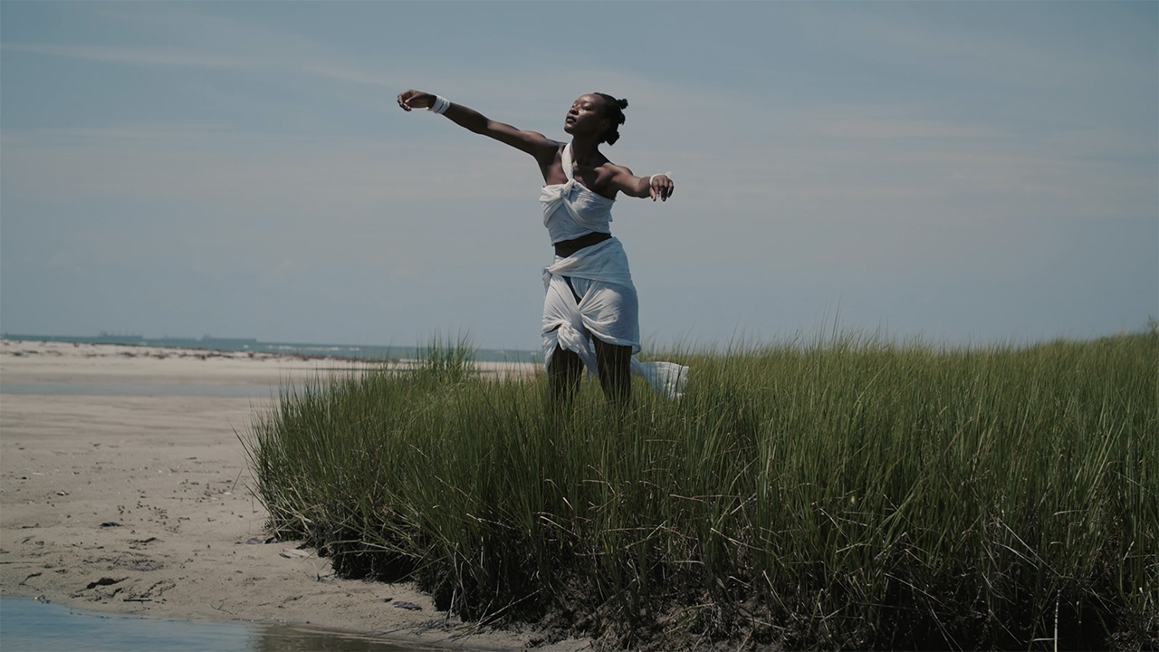 A dancer stands in tall grass along a shoreline. They wear a crop top and skirt made out of knotted white fabric and their hair forms three buns down the back of their head. They stand, extending their arms outwards, as sunlight hits their face.