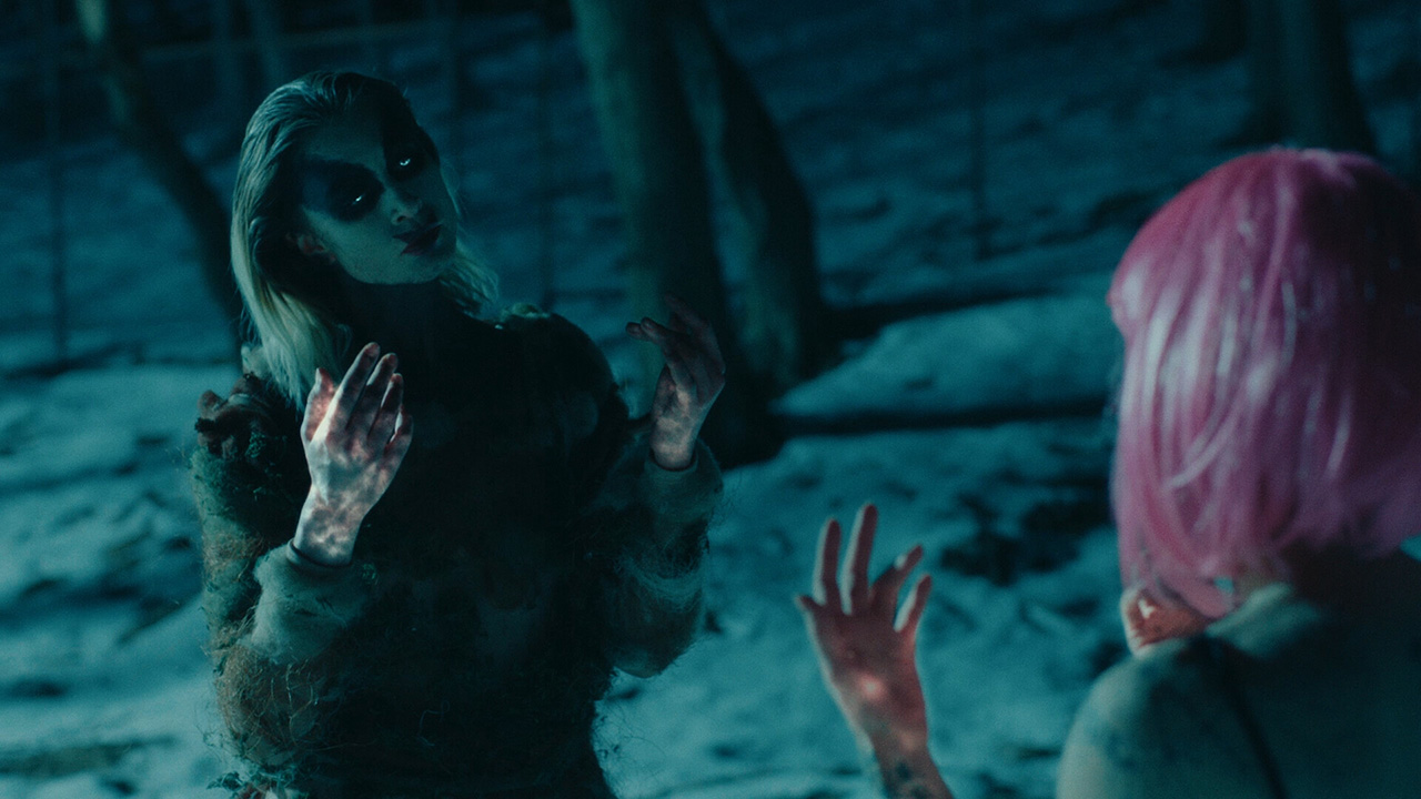 In a dark snowy landscape, two figures mirror each other with their hands raised. One has a short, bubblegum pink bob. The other looks otherworldly with a pale face, dark markings around their glowing eyes, and tattered clothing. Their hands glow dimly.