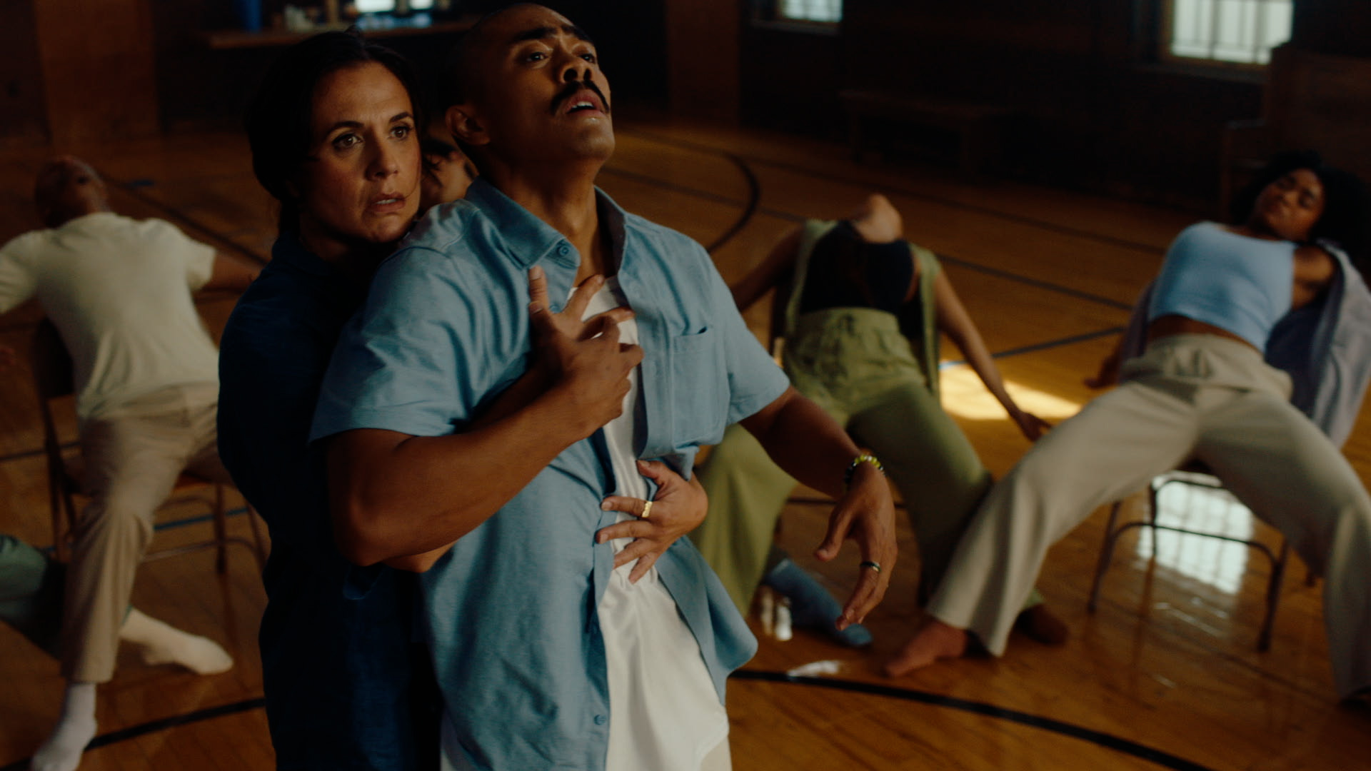 Two dancers look intently at something off camera as one clutches the other. They wear loose blue shirts and stand on the wooden floor of a basketball gym. In a semi-circle around them, dancers sit in splayed positions in metal folding chairs.