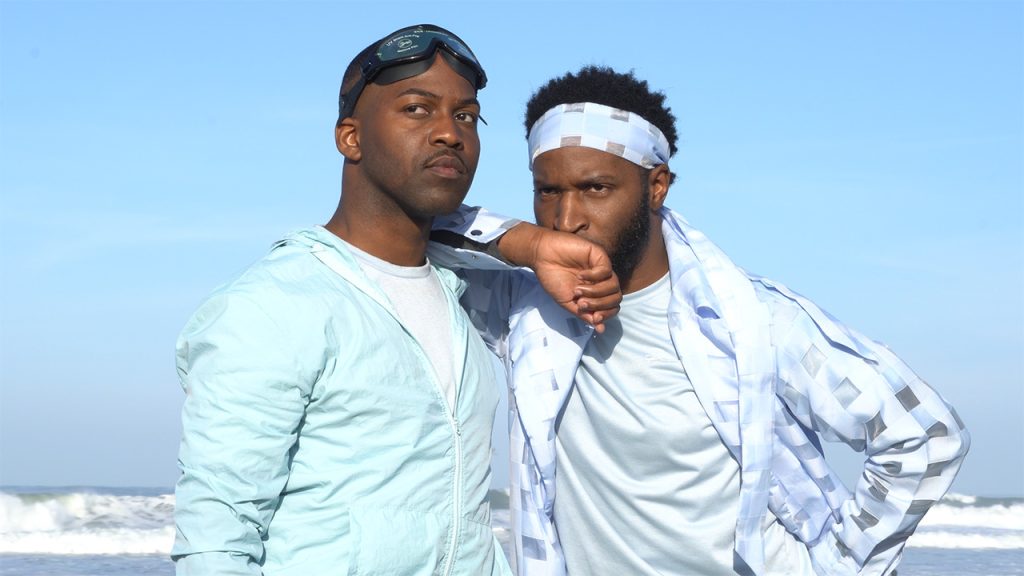 Two people wear bright blue clothing that matches the clear sky behind them. One rests his arm on the other's shoulders, and they look intently towards the same spot in the distance. One of them wears swim goggles pushed up to his forehead.