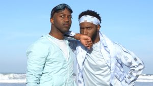 Two people wear bright blue clothing that matches the clear sky behind them. One rests his arm on the other's shoulders, and they look intently towards the same spot in the distance. One of them wears swim goggles pushed up to his forehead.