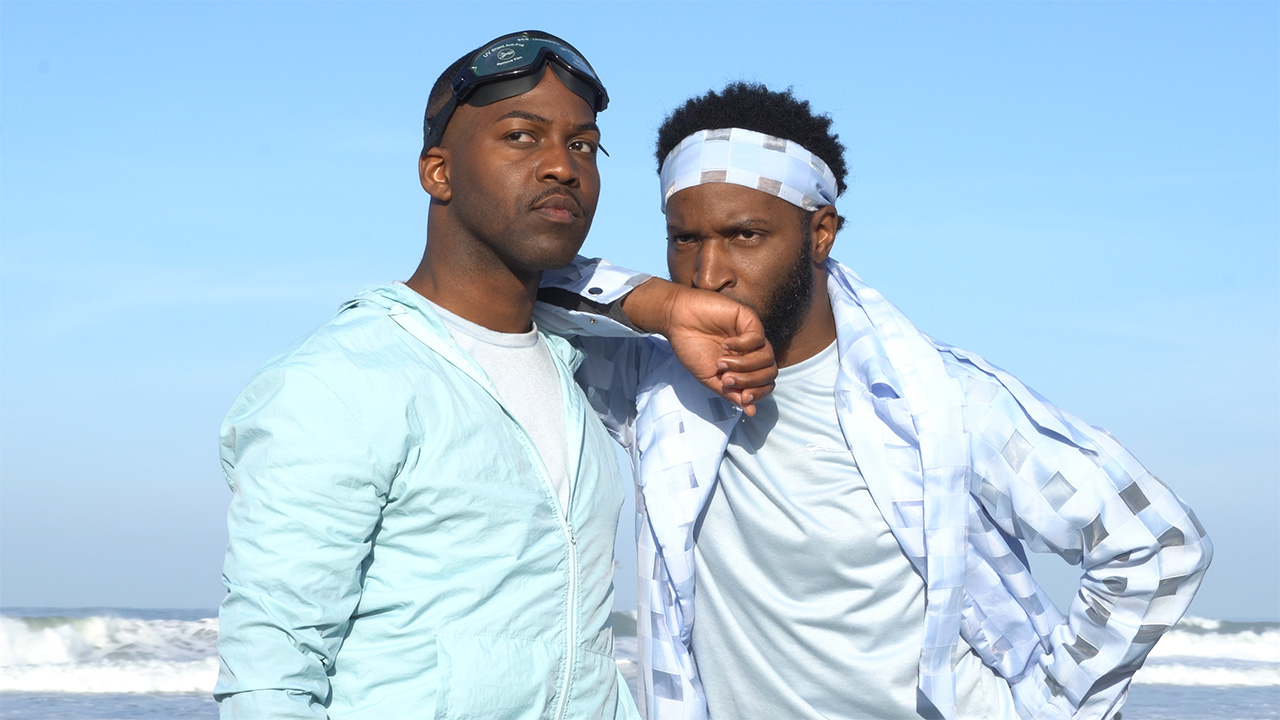 Two people wear bright blue clothing that matches the clear sky behind them. One rests his arm on the other's shoulders, and they look intently towards the same spot in the distance. One of them wears swim goggles pushed up to his forehead.