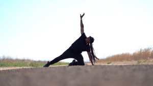 A black dancer with long locks in a ponytail kneels with one knee and a hand resting on the ground, and the other leg extending out to the side. His other arm reaches towards the sky, and he grist his teeth, eyes squinting. He wears blacks jeans, a black t-shirt and black beanie.