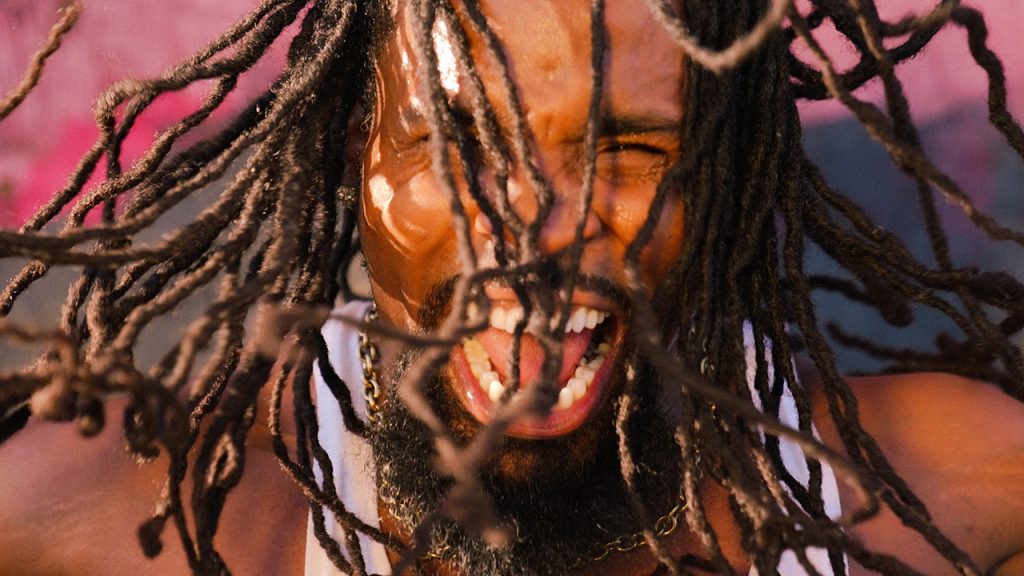Andrew "3D Dance Fever" Jones yells into the camera with lots of emotion, his hair flying around his face. He is a black man wearing a white sleeveless shirt and stands in front of a pink wall.