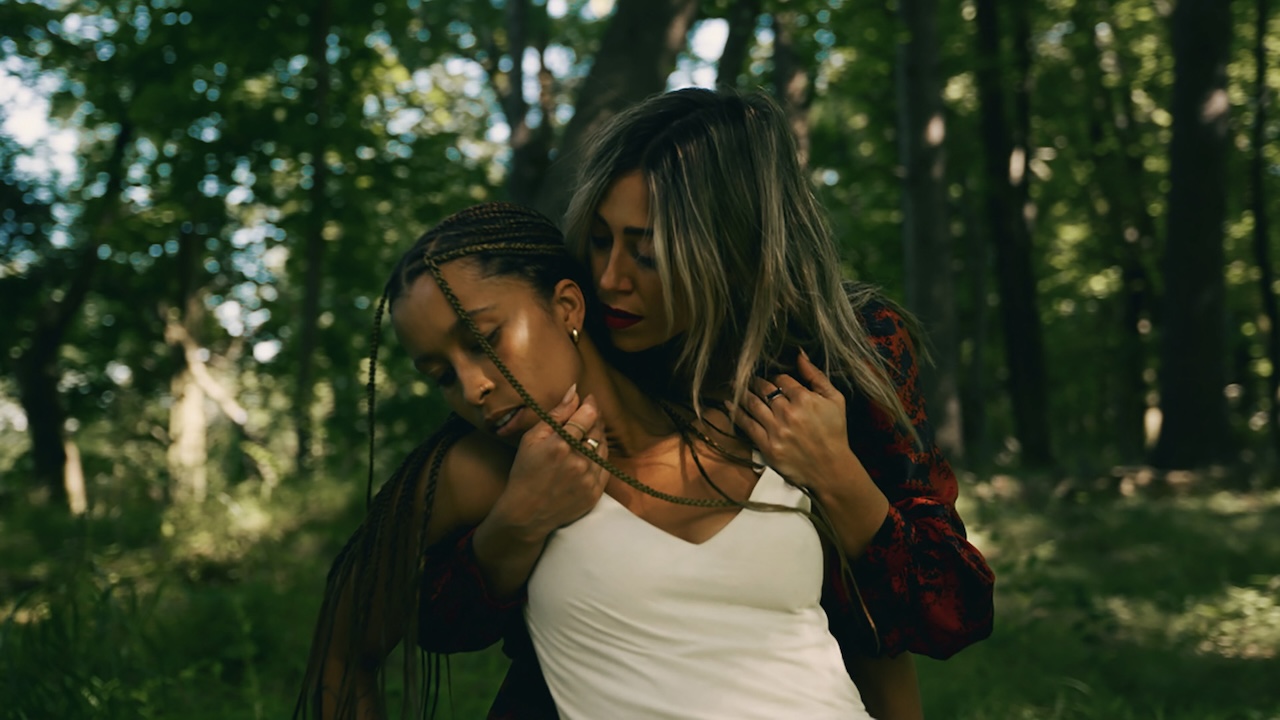 As the sunlight streams through the branches of the foreest, a woman with dark-rooted bleached hair embraces another woman with braids in a white tank top from behind.
