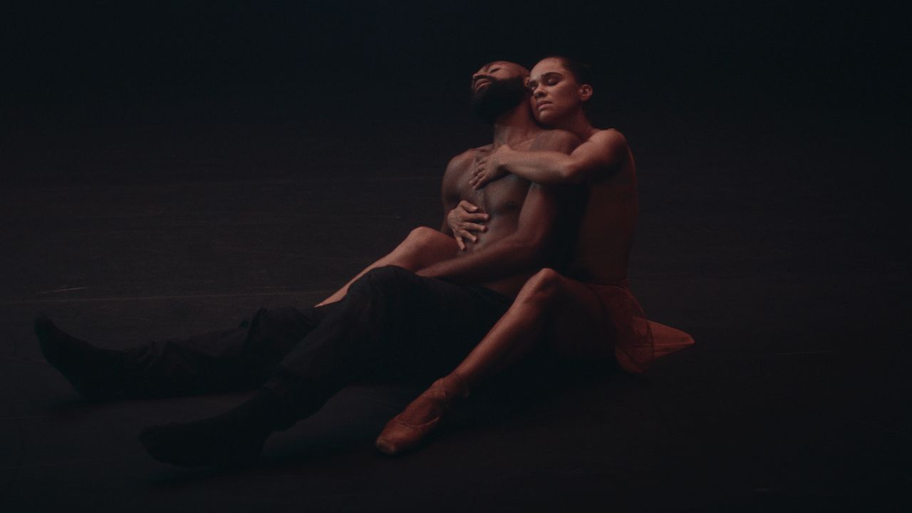 Misty Copeland and Babtunji Johnson sit on a dark floor. Misty sits behind Babatunji, her arms holding his bare chest. He leans his head back on her shoulder.