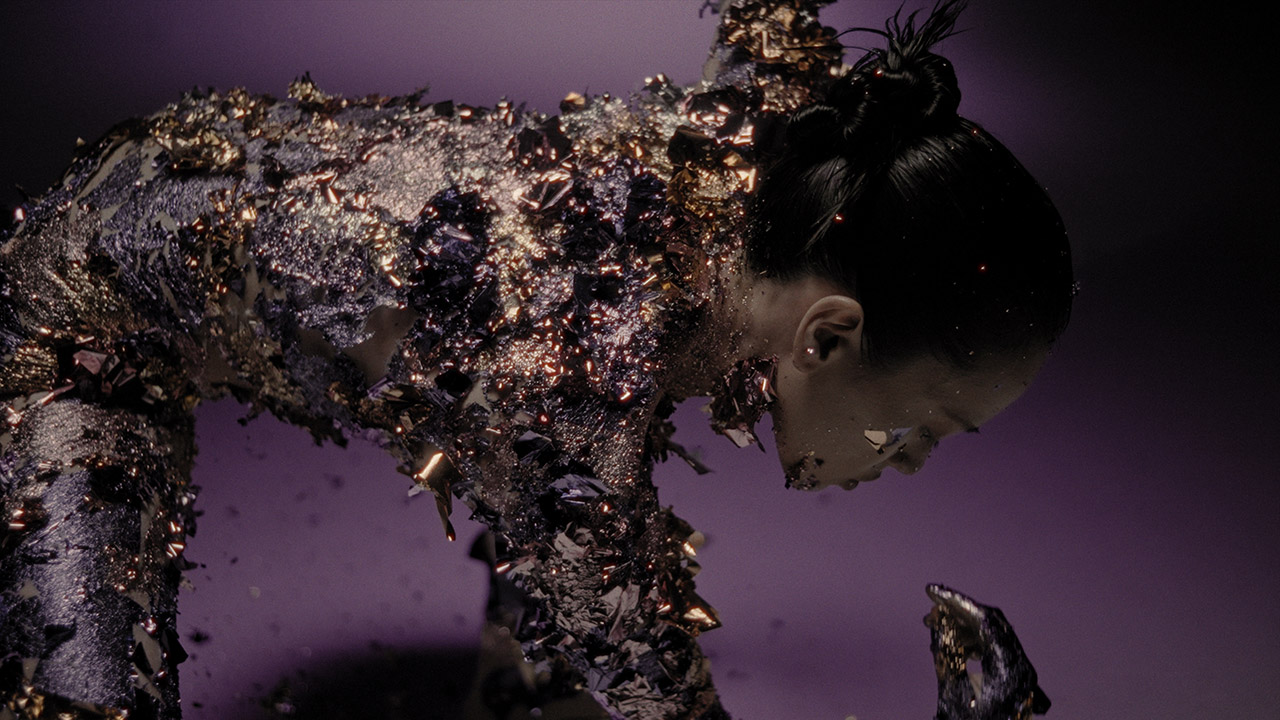 An Asian woman with a slick bun hairstyle crouches over, as her entire body is covered in black ash and metallic gold leaf.