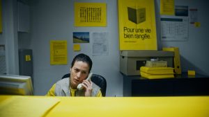 An office worker sits at a desk with a corded phone up to their ear. They wear a yellow shirt under a gray vest, and their face shows annoyance. There are splashes of the same shade of yellow all around the office.