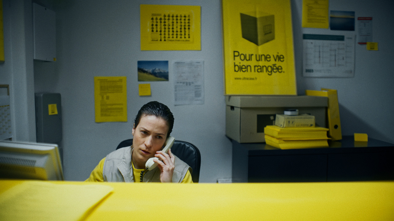 An office worker sits at a desk with a corded phone up to their ear. They wear a yellow shirt under a gray vest, and their face shows annoyance. There are splashes of the same shade of yellow all around the office.