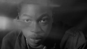 A greyscale image of a young black person staring intensely into the camera lens.