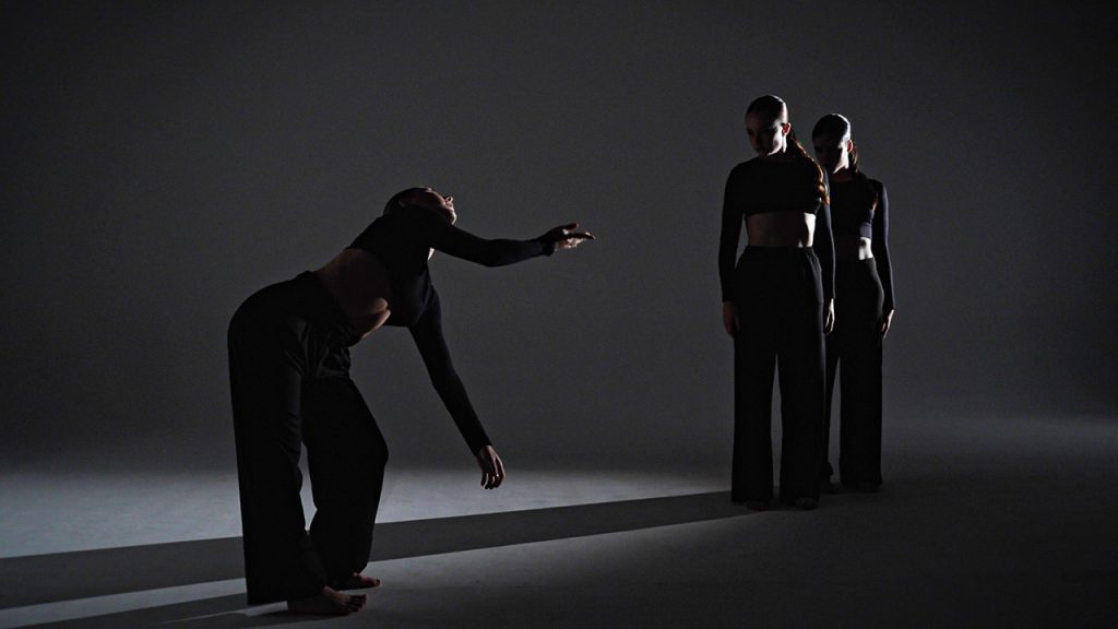 Three dancers stand in a dark photo studio, and a light shines on them from the side, causing a long shadow across the floor. Two of the dancers stand close together, looking at one who is bowed at the waist with one arm reaching out towards the other dancers and another arm dangling below their chest.