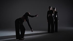 Three dancers stand in a dark photo studio, and a light shines on them from the side, causing a long shadow across the floor. Two of the dancers stand close together, looking at one who is bowed at the waist with one arm reaching out towards the other dancers and another arm dangling below their chest.
