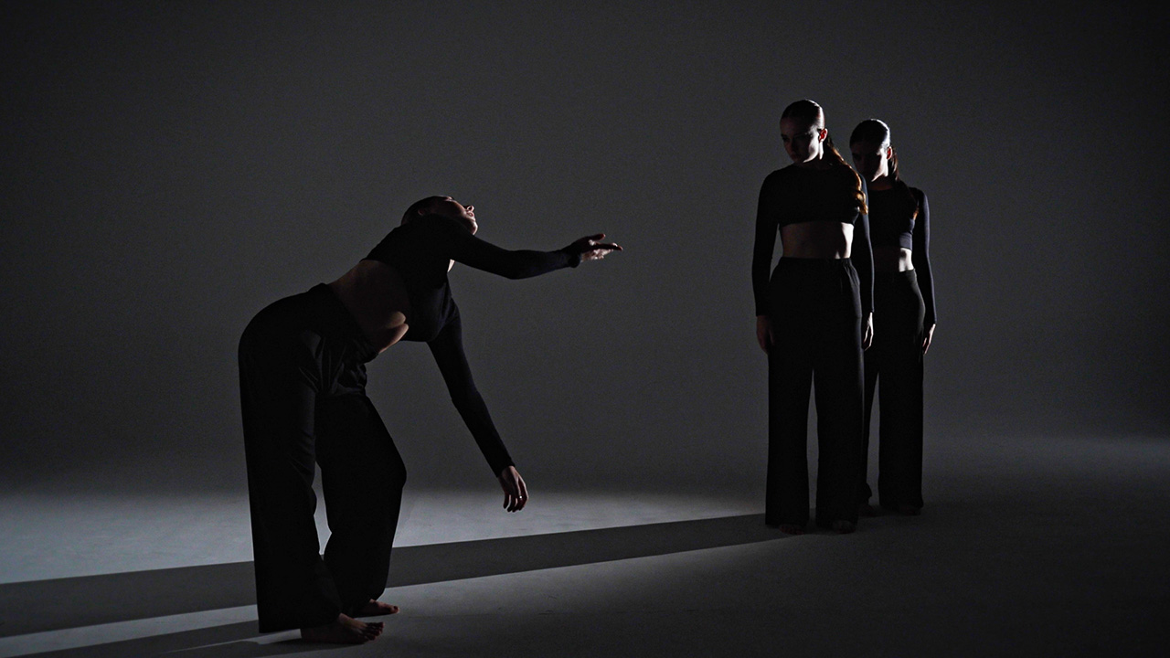 Three dancers stand in a dark photo studio, and a light shines on them from the side, causing a long shadow across the floor. Two of the dancers stand close together, looking at one who is bowed at the waist with one arm reaching out towards the other dancers and another arm dangling below their chest.