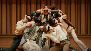A group of nine dancers in sunglasses cluster around one seated in a chair with orange sunglasses and long nails. They all wear vintage 70s clothes in earth tones and are indoors on a wooden stage.