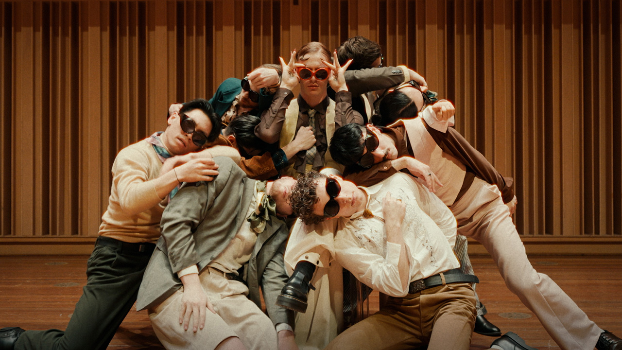 A group of nine dancers in sunglasses cluster around one seated in a chair with orange sunglasses and long nails. They all wear vintage 70s clothes in earth tones and are indoors on a wooden stage.