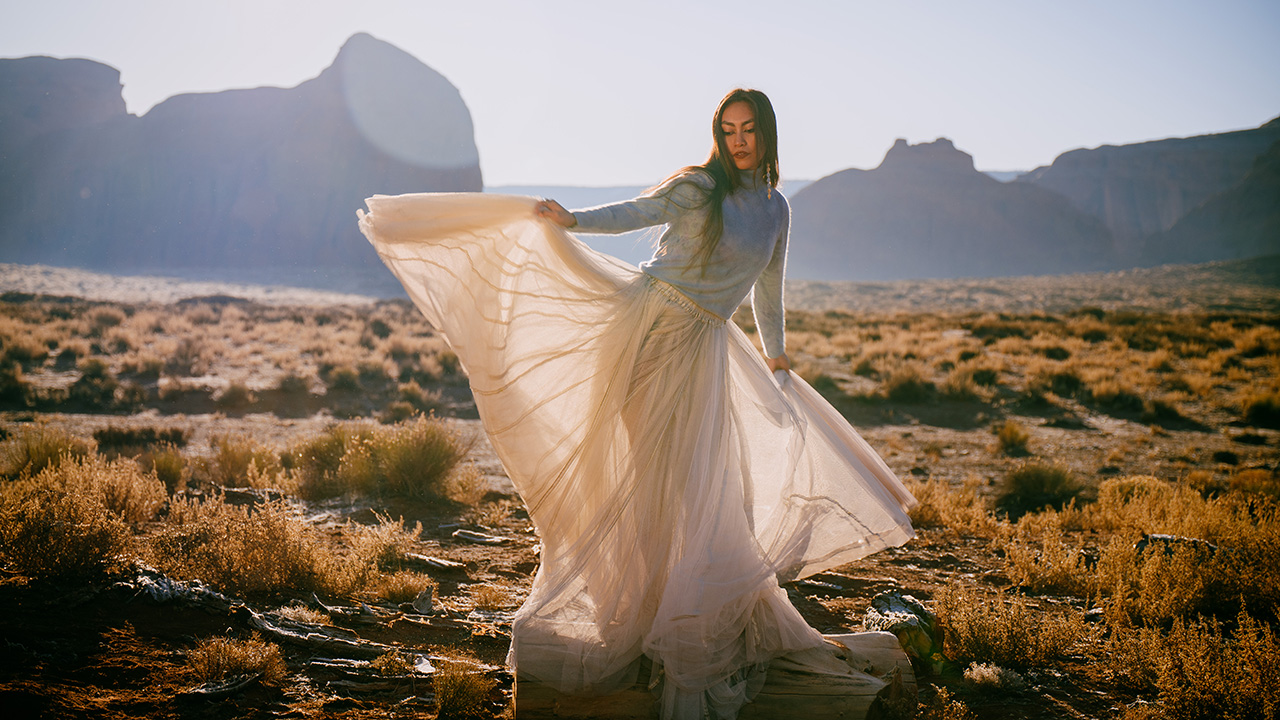 Than Povi Martinez, a Native American dancer, stands in a sunny desert. She wears a thin, beige dress, holding it up by her sides. Light shines gently through it. Her long, dark hair falls over one shoulder.