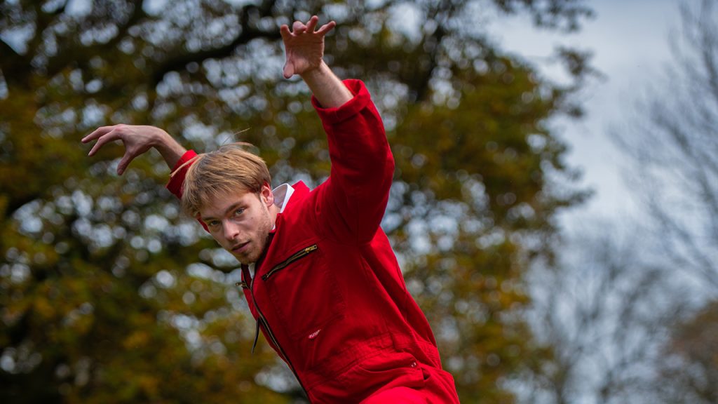A blonde dancer with blue eyes and stubble wears a bright red jumpsuit with a white collared shirt underneath. He leans slightly forward with his arms above and behind his head, and with his hands in claw shapes. He looks directly at the camera.